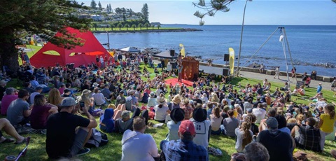 Crowd of people sitting on grass at festival watching performers in large red tent, overlooking Kiama Harbour